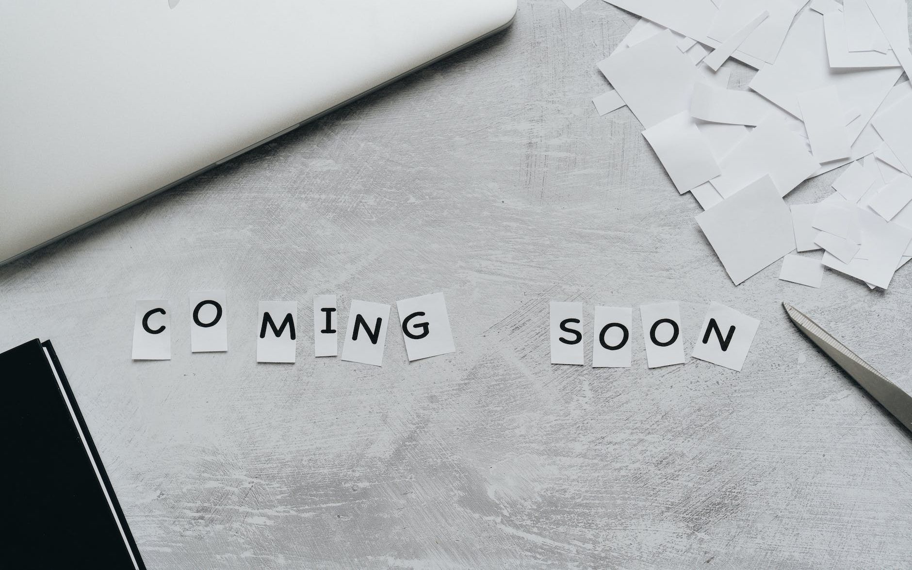 "Coming soon" written in letters cut out of paper. Cut up paper and scissors are also visible along with the edges of a notebook and laptp. 
