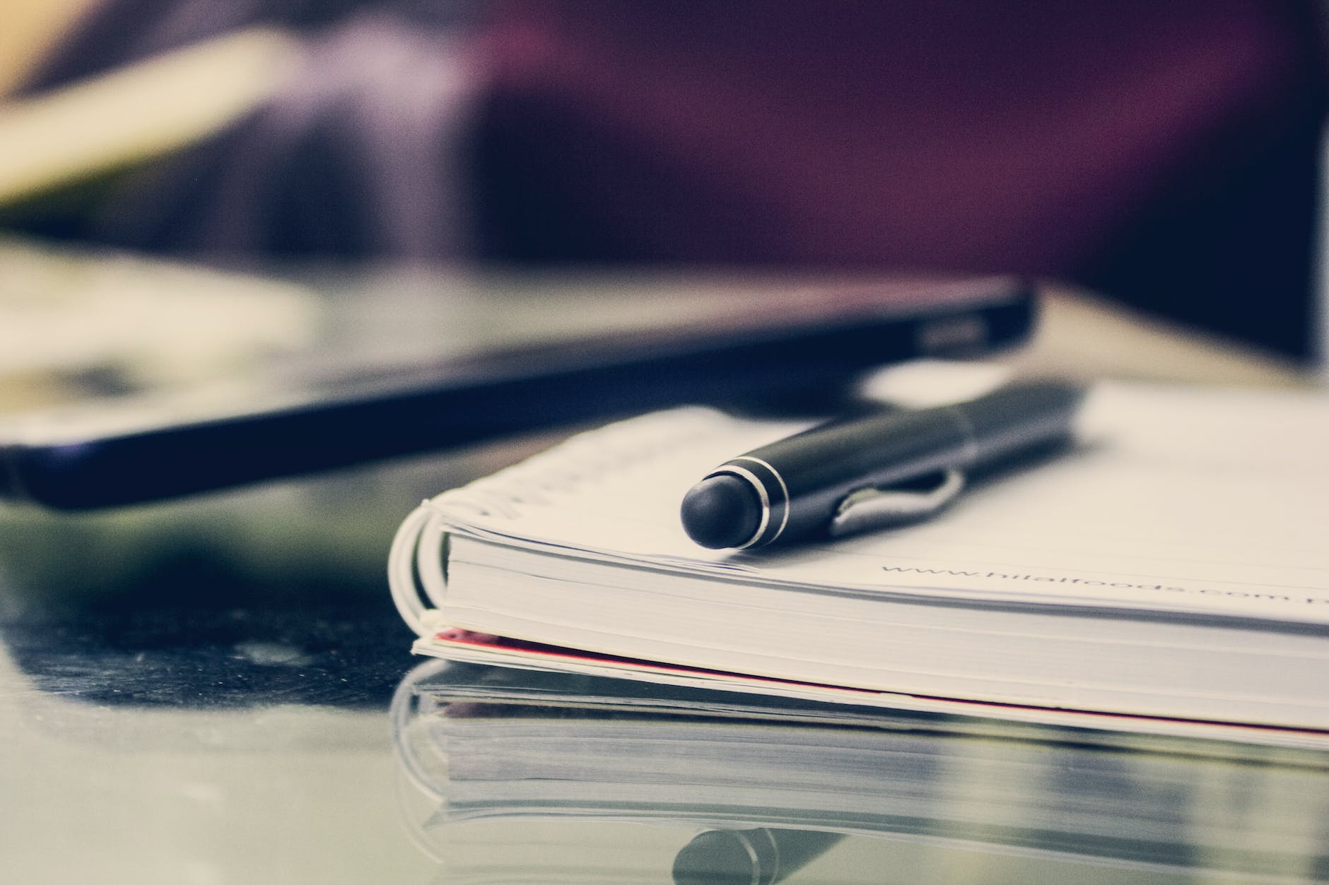 A pen lying on top of a notebook. A phone is lying next to the notebook, but the image is out of focus. 